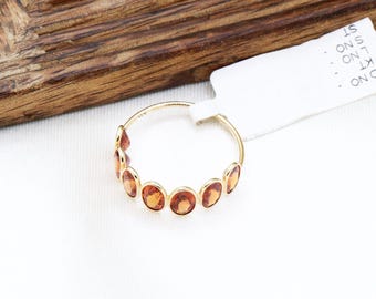 May include: A gold ring with a row of seven orange gemstones. The ring is on a white surface.