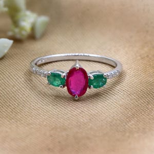 May include: A silver ring featuring a large oval-cut ruby center stone flanked by two smaller emeralds. The band is adorned with small, clear accent stones. The ring is set against a neutral, textured background.
