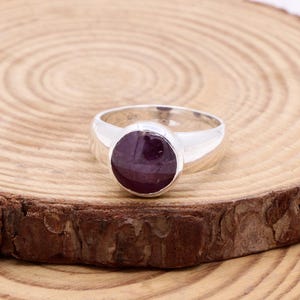 May include: A silver ring featuring a large, circular, deep purple gemstone. The ring is presented on a wooden surface with a natural, rustic appearance. The ring's band is smooth and polished. The gemstone is set in a bezel.