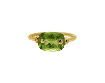 May include: A gold ring featuring a green gemstone. The gemstone is set in a bezel setting and has a cushion cut. The ring is crafted from gold wire and has two small gold beads on either side of the gemstone.