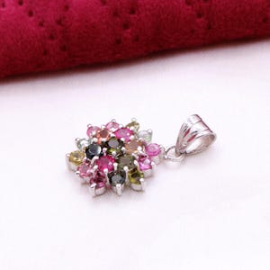 May include: A silver pendant featuring a cluster of colorful gemstones in shades of pink, green, yellow, and black. The pendant has a floral design and is set against a white background with a red fabric in the background.