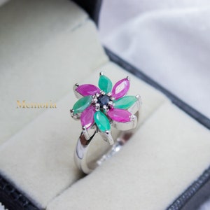 May include: A silver ring with a flower design featuring pink and green gemstones. The center of the flower has a small blue gemstone.