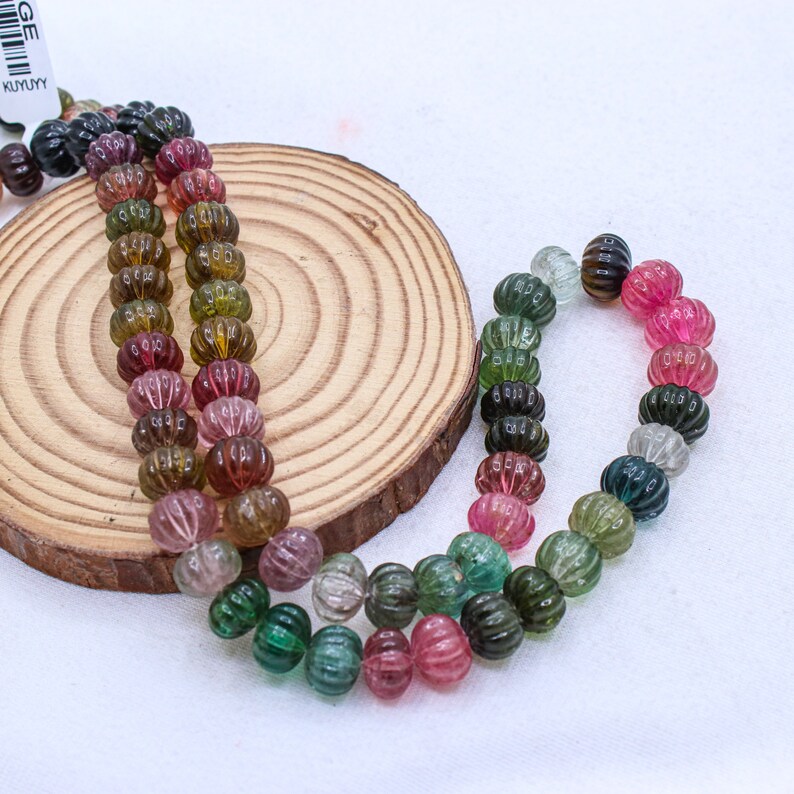 May include: A strand of multi-colored watermelon tourmaline beads. The beads are faceted and have a ribbed texture. The beads are arranged in a rainbow pattern, with pink, green, and black beads.