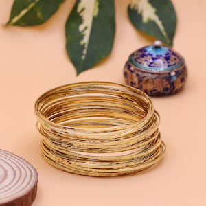 May include: A stack of 14 gold-toned bangles with a hammered texture. The bangles are stacked on a light pink surface.