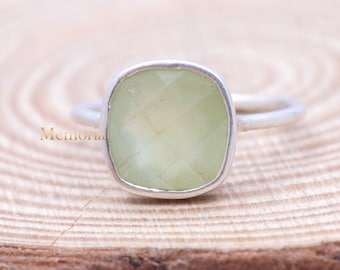 May include: A silver ring with a light green square-shaped gemstone. The ring is on a wooden surface.