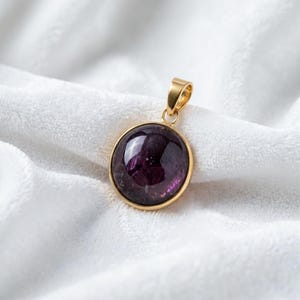May include: A round pendant featuring a deep purple, translucent gemstone set in a polished gold-toned metal frame. The pendant includes a small, matching gold-toned bail at the top for attachment, showcasing a smooth, domed surface.