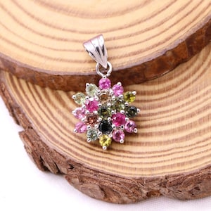 May include: A silver pendant featuring a flower design made of colorful gemstones. The gemstones are various shades of pink, green, and black. The pendant is displayed on a wooden surface.