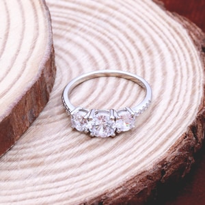 Könnte beinhalten: Ein silberner Ring mit drei runden, klaren Steinen, die in einer Reihe angeordnet sind. Der Ring liegt auf einer Holzoberfläche.