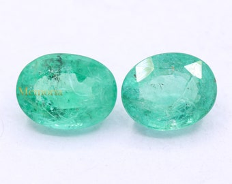 May include: Two oval-shaped emerald gemstones, each with a green hue and a slightly different shade. The gemstones are set against a white background.