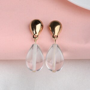 Może przedstawiać: Para złotych kolczyków wiszących z przezroczystymi, łzowymi koralikami szklanymi.