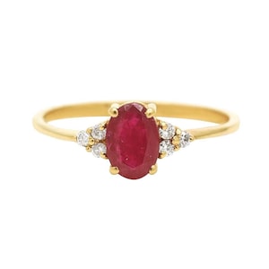 May include: A gold ring featuring a central oval-cut red ruby gemstone held by four prongs. The ruby is flanked on each side by three small, round, clear white diamonds set in a cluster, all mounted on a thin, polished yellow gold band.