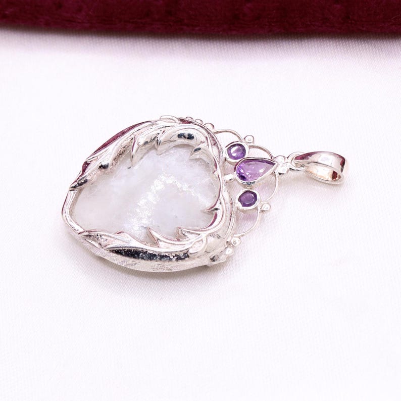 Peut inclure: Pendentif en argent avec une pierre blanche iris&eacute;e en forme de c&oelig;ur, encadr&eacute;e d'une monture argent&eacute;e d&eacute;corative. Le pendentif comporte trois petites pierres pr&eacute;cieuses violettes et une boucle pour une cha&icirc;ne. Design orn&eacute; et &eacute;l&eacute;gant.