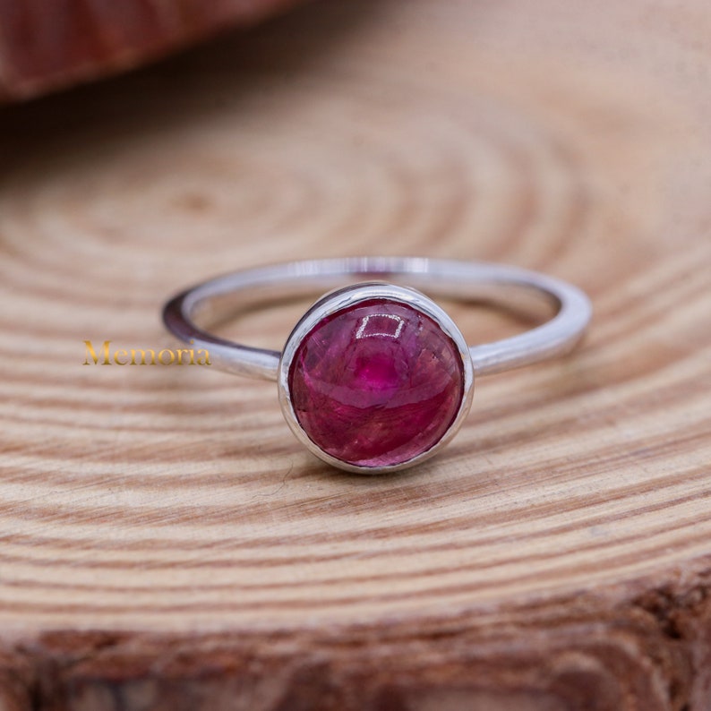 Puede incluir: Un anillo de plata con una piedra preciosa redonda de color rosa engastada en un engaste de bisel. El anillo est&aacute; sobre una superficie de madera.