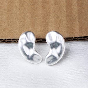 以下が含まれることがあります： 銀色の豆の形をしたイヤリングのペア。イヤリングは滑らかで、わずかにテクスチャ加工された表面をしています。