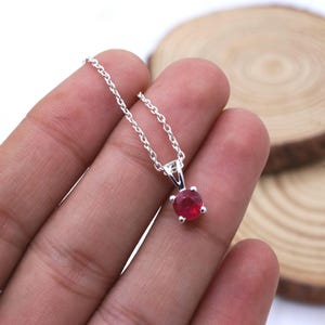 May include: A silver pendant necklace with a round, faceted ruby gemstone. The ruby is a vibrant red colour and is set in a four-prong setting. The chain is a delicate silver chain.