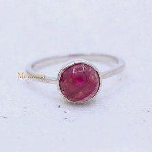 Puede incluir: Un anillo de plata con una piedra preciosa rosa. La piedra es redonda y tiene una superficie lisa y pulida.