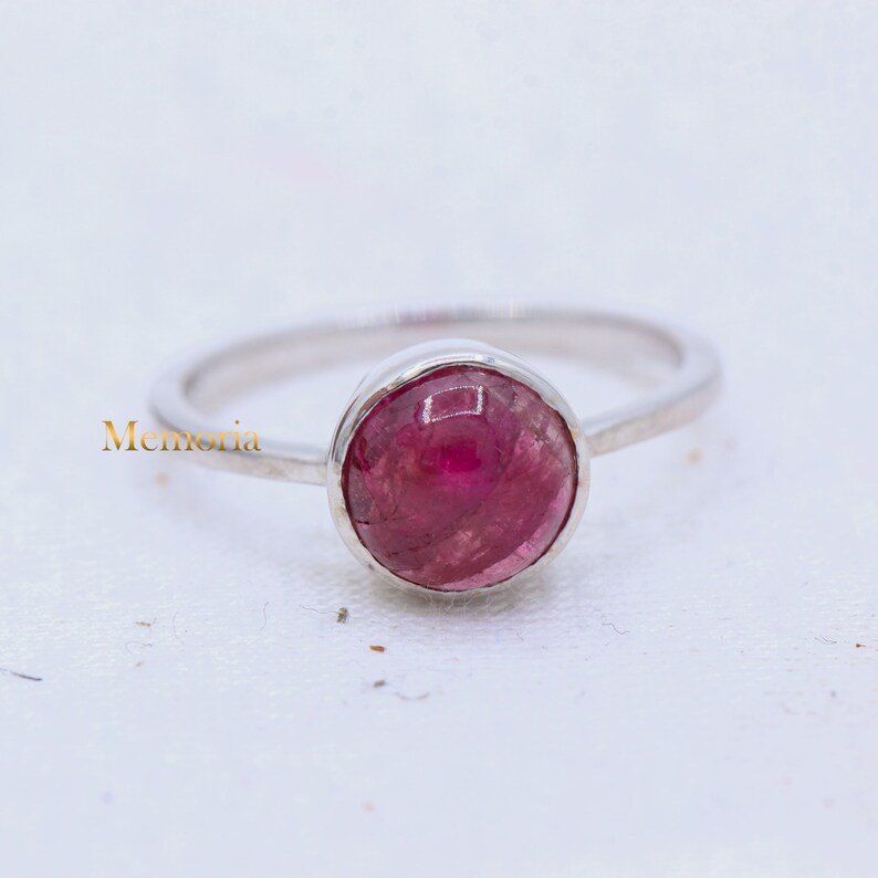 Puede incluir: Un anillo de plata con una piedra preciosa rosa. La piedra es redonda y tiene una superficie lisa y pulida.