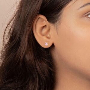 Close-up of a person's ear wearing a small square pink gemstone stud earring in the earlobe.