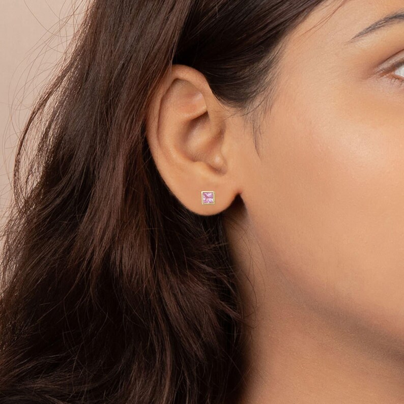 Close-up of a person's ear wearing a small square pink gemstone stud earring in the earlobe.
