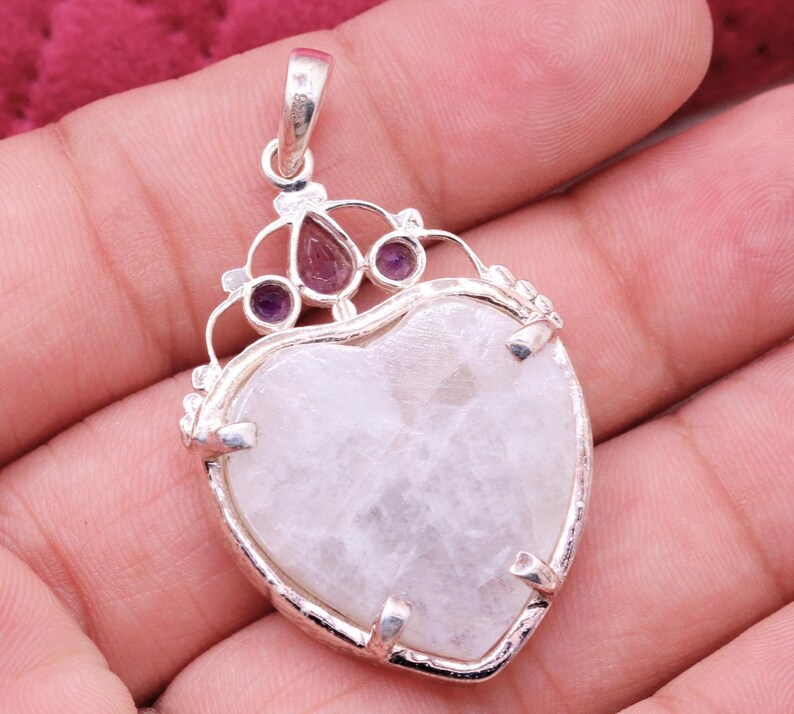 Peut inclure: Pendentif en forme de c&oelig;ur avec un cadre argent&eacute;. Le pendentif pr&eacute;sente une grosse pierre claire au centre, avec de plus petites pierres violettes et rouges en haut. Le pendentif est orn&eacute; d'un &eacute;l&eacute;ment d&eacute;coratif en forme de couronne.