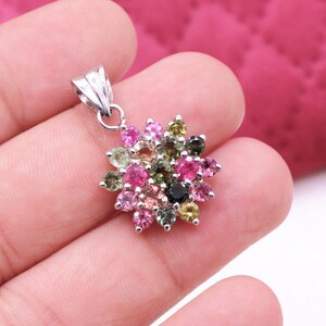 May include: A silver pendant featuring a flower design. The pendant is adorned with a cluster of colorful gemstones in shades of pink, green, and black. The pendant is held in a hand.