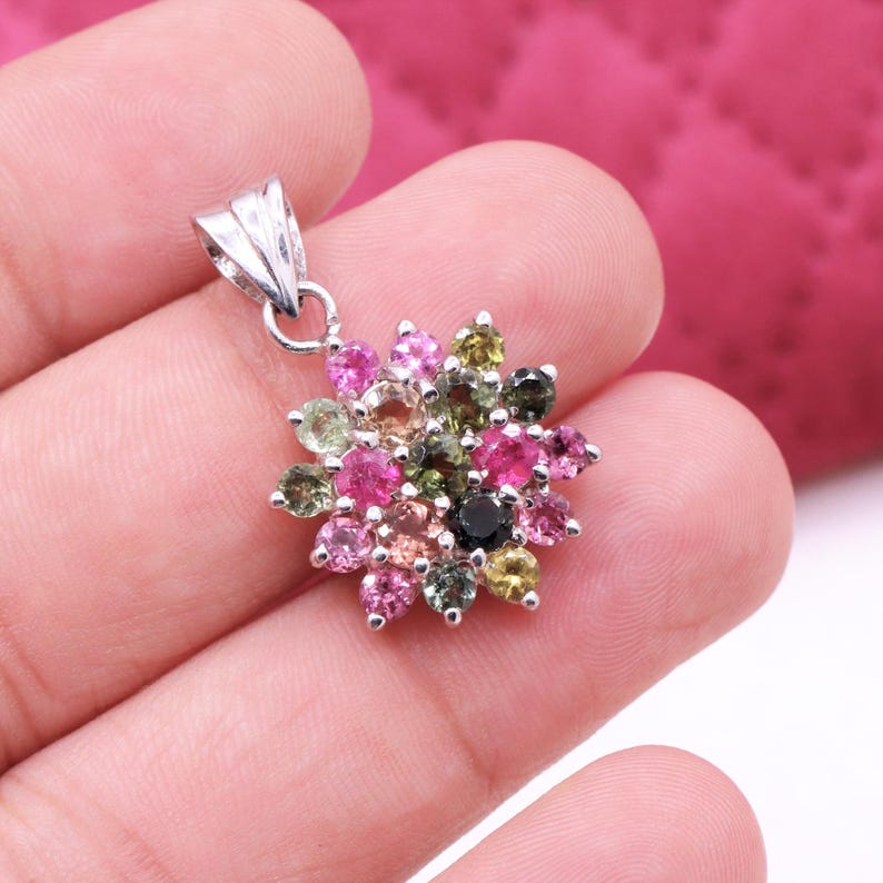May include: A silver pendant featuring a flower design. The pendant is adorned with a cluster of colorful gemstones in shades of pink, green, and black. The pendant is held in a hand.