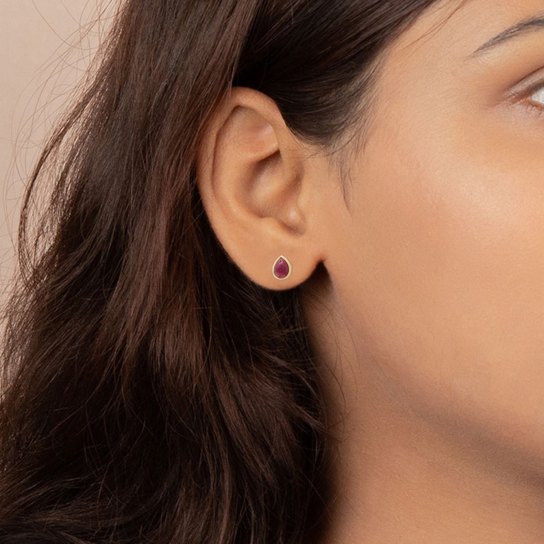 May include: A teardrop-shaped stud earring featuring a deep pink gemstone set in a smooth gold-toned metal bezel. The earring is positioned on the earlobe, showcasing a minimalist design with a polished finish and a secure, simple backing.