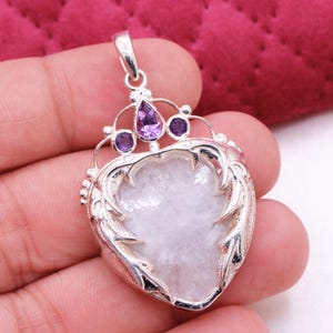 Peut inclure: Un pendentif en argent avec une pierre en forme de c&oelig;ur de couleur claire. Le pendentif pr&eacute;sente un motif de couronne avec trois pierres pr&eacute;cieuses violettes. Le pendentif est tenu dans une main.