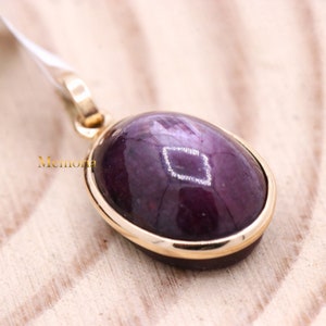 Puede incluir: Un colgante de oro con una piedra ovalada de color púrpura oscuro engastada en un bisel de oro. La piedra tiene un brillo sutil.