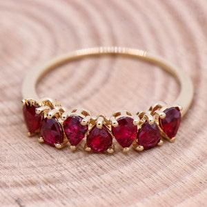 May include: A gold ring featuring seven vibrant red pear-shaped gemstones. The gemstones are set closely together, creating a continuous row of color. The ring is displayed on a light-colored wooden surface.