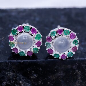 Puede incluir: Pendientes de botón con una piedra central clara rodeada por un halo de gemas verdes y magenta alternas. Los pendientes tienen un engaste de metal plateado y se muestran sobre una superficie oscura. Son una joya.