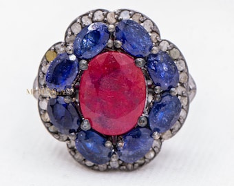 May include: A silver ring featuring a large red oval gemstone surrounded by smaller blue oval gemstones and smaller clear round gemstones.