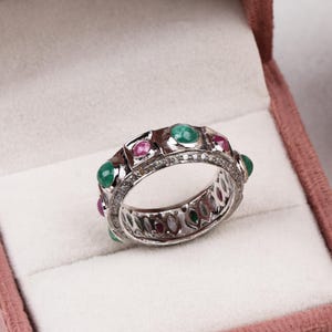 Puede incluir: Un anillo de plata con piedras preciosas de esmeralda y rubí alternas, acentuado con pequeños diamantes. El anillo está engastado en una banda con un diseño detallado y se muestra en una caja de joyería rosa.