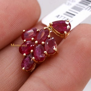 May include: A gold pendant featuring a cluster of six oval-cut deep red rubies arranged in a floral pattern, with a single pear-cut ruby attached at the top. The word Memoria is printed in gold on the left side of the ruby cluster.