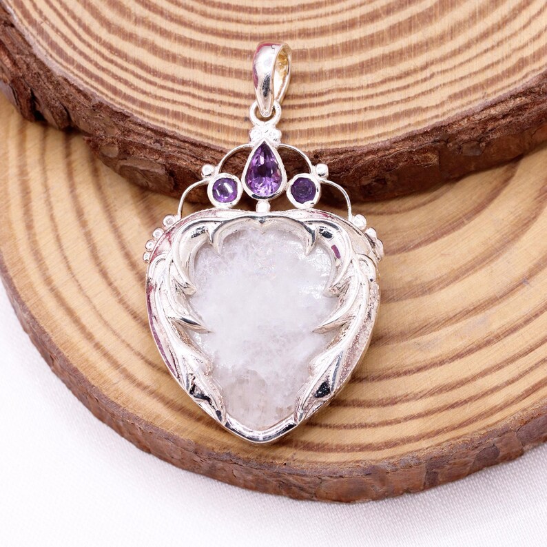 Peut inclure: Pendentif en argent avec une pierre blanche en forme de c&oelig;ur. Le pendentif est orn&eacute; de pierres pr&eacute;cieuses violettes et de filigranes argent&eacute;s d&eacute;coratifs. La b&eacute;li&egrave;re est dor&eacute;e. Le pendentif est pr&eacute;sent&eacute; sur une surface en bois.
