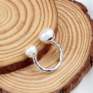 May include: A silver ring with two white pearl accents. The ring is a simple, open circle design.