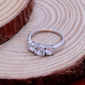 May include: A silver ring with three round clear stones set in a row. The ring is on a wooden surface.