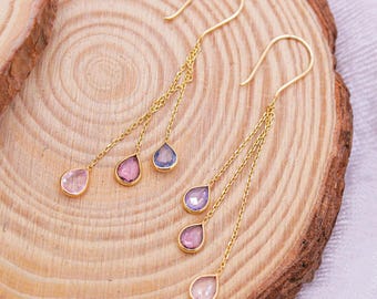 May include: Gold dangle earrings with three teardrop-shaped gemstones. The gemstones are pink, purple, and clear.