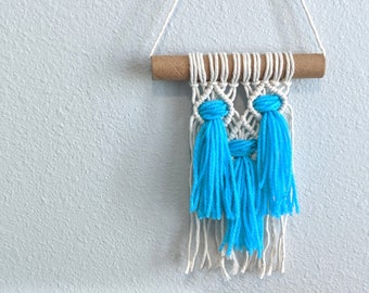 Pode incluir: Uma tapeçaria de macramé branca com borlas azuis. A tapeçaria está pendurada em um cordão branco e está presa a uma vareta de madeira. A tapeçaria está na frente de um quadro de letras azul claro com o texto "Make each day your masterpiece".