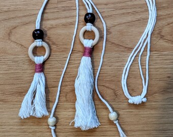 Pode incluir: Três colares de cordão de algodão branco com contas de madeira e borlas. Cada colar tem um anel de madeira com uma borla de algodão branco e uma conta de madeira castanho. As borlas são de cor rosa claro.