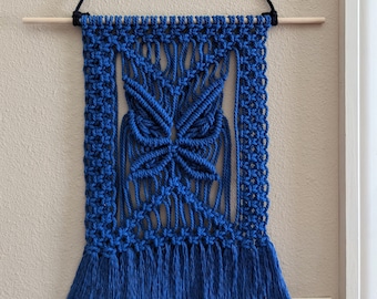 Pode incluir: Decoração de parede em macramé azul com um design de borboleta e franjas. A decoração é presa a uma haste de madeira e tem um suporte de corda preto.