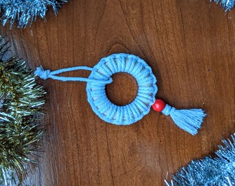 Pode incluir: Um ornamento de macramé azul claro com uma conta vermelha e uma borla pendurada em uma superfície de madeira cercada por serpentina azul e dourada.