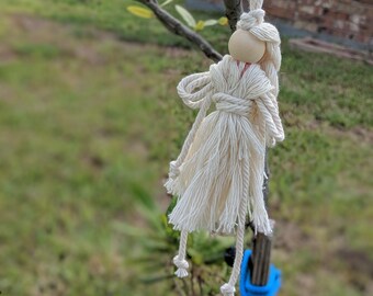 Pode incluir: Uma boneca de fio branco com cabeça de madeira pendurada em um galho de árvore. A boneca tem uma saia longa e fluida e uma cintura amarrada.