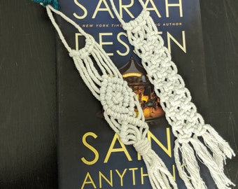 Pode incluir: Dois marcadores de livro de macramé branco com borlas. Os marcadores estão em cima de um livro com o título "Sarah dessen,  San anything" na capa.