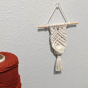 Pode incluir: Uma tapeçaria de macramé branca com uma vareta de madeira e uma borla branca. A tapeçaria tem um pequeno objeto branco no centro.