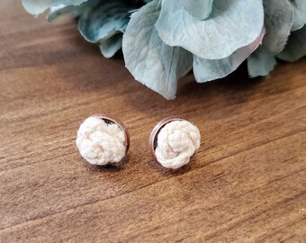 Pode incluir: Um par de brincos de nó de corda branca com suporte marrom. Os brincos estão sobre uma superfície de madeira.