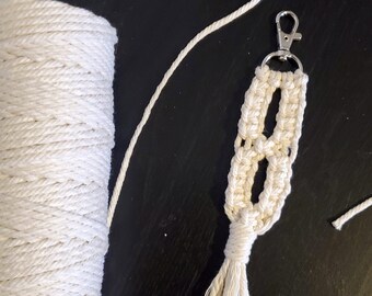 Pode incluir: Um chaveiro de macramé branco com um chaveiro prateado e uma borla. O chaveiro é feito com um cordão branco grosso e tem um design simples. Também há um carretel de cordão branco na imagem.
