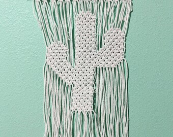 Pode incluir: Decoração de parede de macramé branca em forma de cacto. O cacto é feito com corda branca e tem uma vareta de madeira na parte superior.