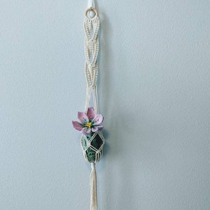 Könnte beinhalten: Ein weißer Makramee-Pflanzenhänger mit einem Holzring oben, der eine kleine grüne Pflanze mit einer einzelnen rosa Blüte hält.