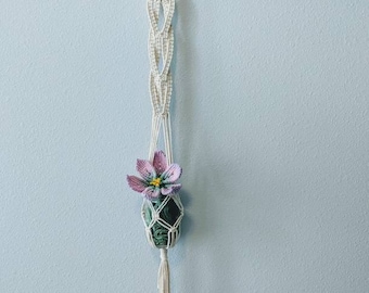 Pode incluir: Um suporte de planta de macramé branco com um anel de madeira no topo, segurando uma pequena planta verde com uma única flor rosa.